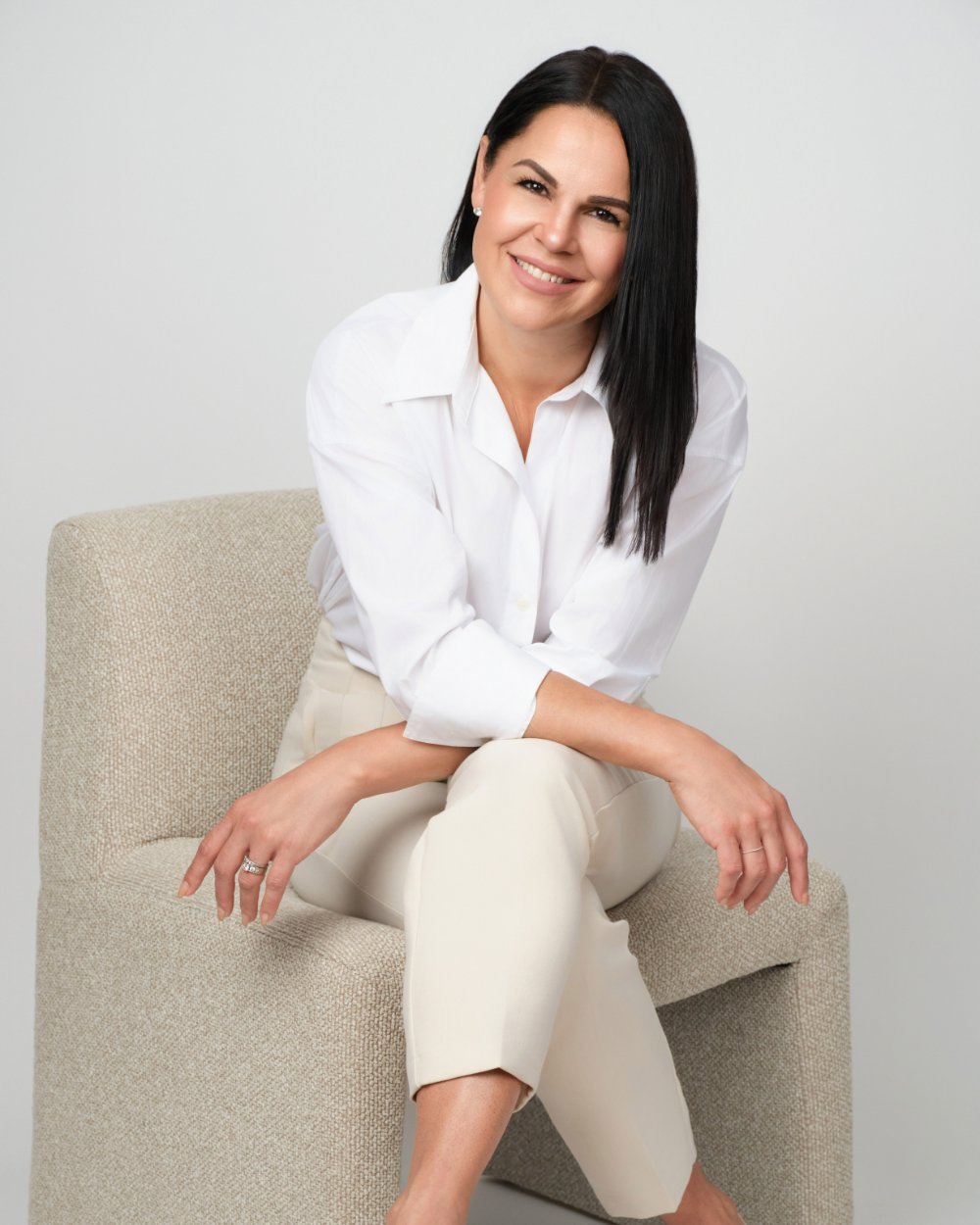Relaxed branding portrait of a woman in a white shirt seated on a modern gray chair in a studio