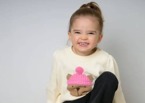 A toddler girl in a cream bear-motif sweater with a scrunched-nose grin