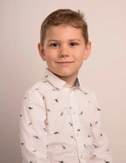 A young boy in a dinosaur-print shirt looking straight to camera on a warm neutral backdrop
