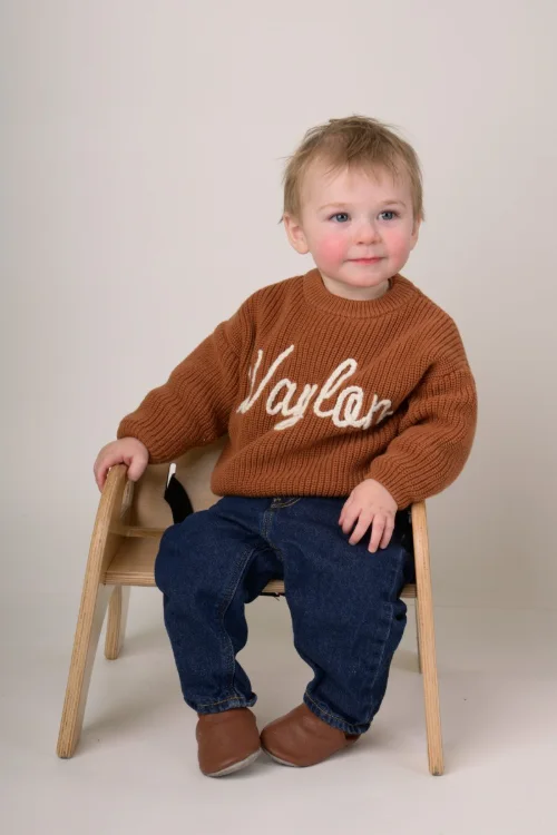 A toddler boy in a rust knit sweater sitting on a small wooden chair