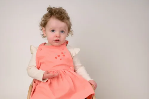 A curly-haired toddler in a coral dress sitting down