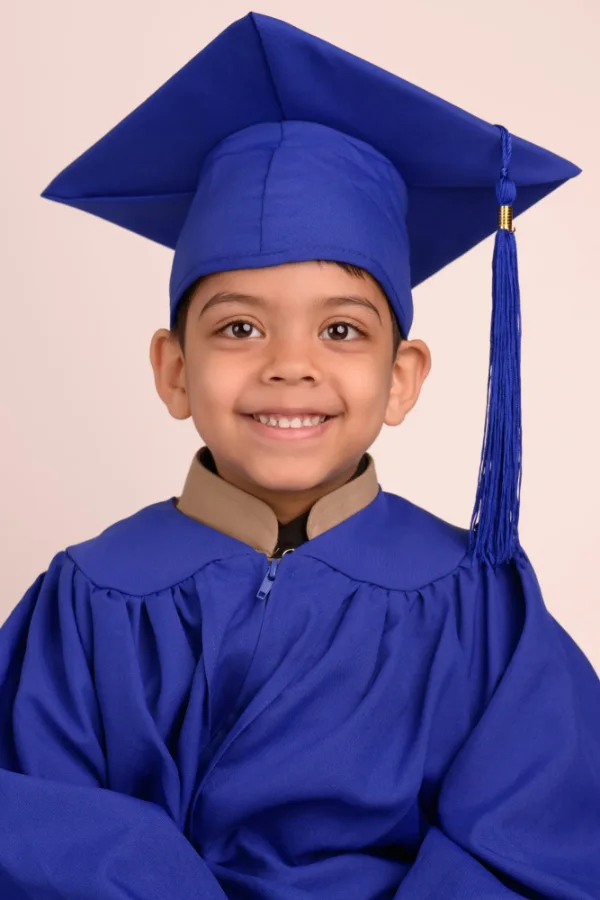 A preschool graduate in a blue cap and gown smiling on a warm blush backdrop