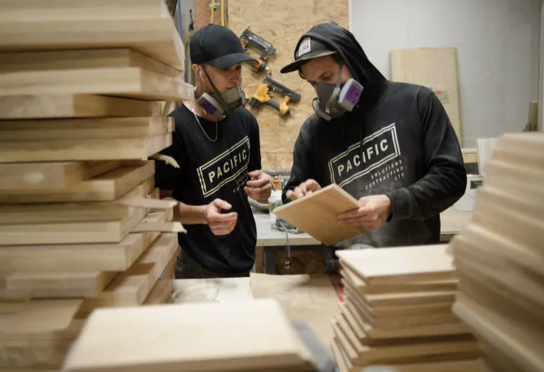 Industrial photography of two millwork tradesmen in respirators inspecting a finished maple panel in a Lower Mainland cabinet shop