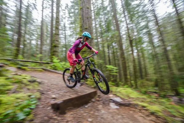 Outdoor brand photography of a mountain biker launching a trail feature with motion-blurred forest behind