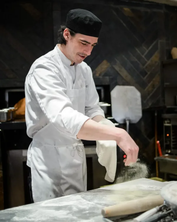Restaurant photography of a chef dusting flour over pizza dough beside a wood-fired oven