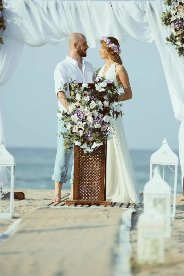 Beach wedding ceremony photographed at a floral podium under a white draped arch at the water’s edge