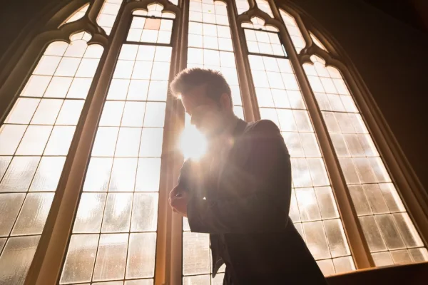 Groom silhouetted in sunlight at a tall church window while fastening his jacket