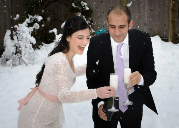 Bride and groom laughing as they toast with champagne in falling snow after their winter wedding