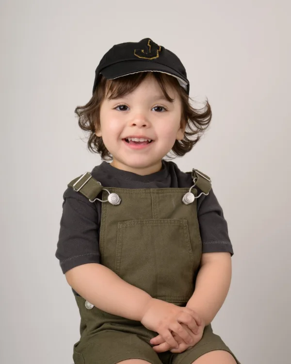 A toddler boy in a black cap and khaki overalls with his hands together