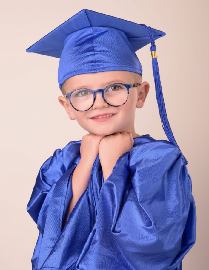 Preschool graduate in a blue cap and gown with his hands tucked under his chin