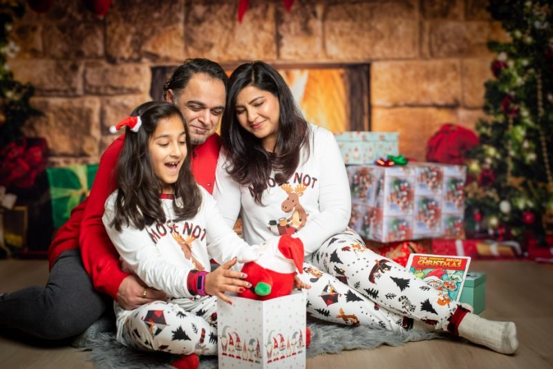Family of three in matching moose pyjamas opening a gift box on the warm fireplace set during a Christmas mini session
