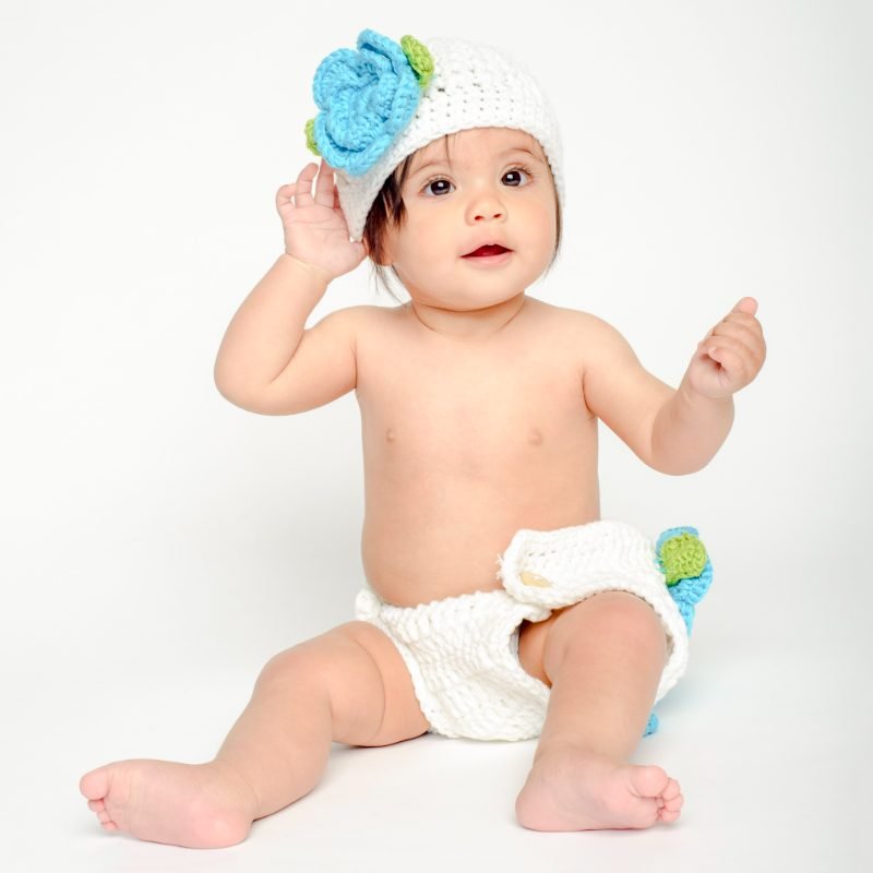 Baby in a white knit hat with a blue flower sitting on a white studio backdrop