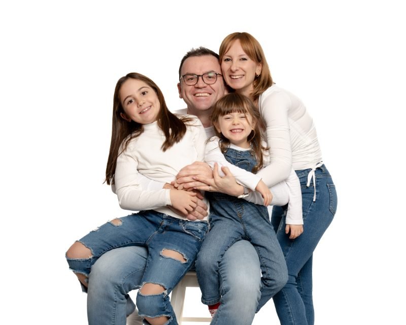 Family of four in white tops and jeans laughing together on a white studio backdrop