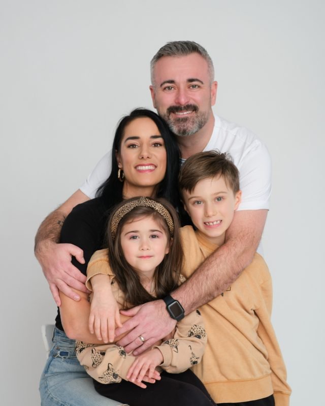 Family of four in a tight hug with the children in tan sweaters on a white studio backdrop