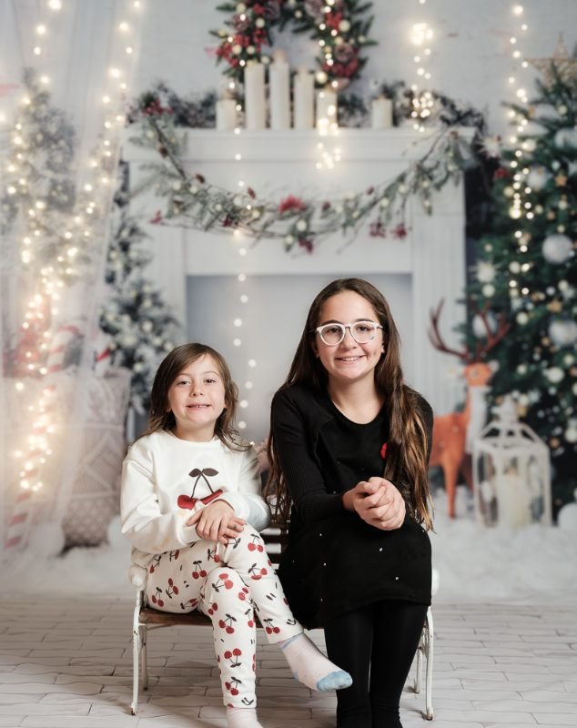Two sisters on a white bench in front of a white fireplace with candles, garland and string lights