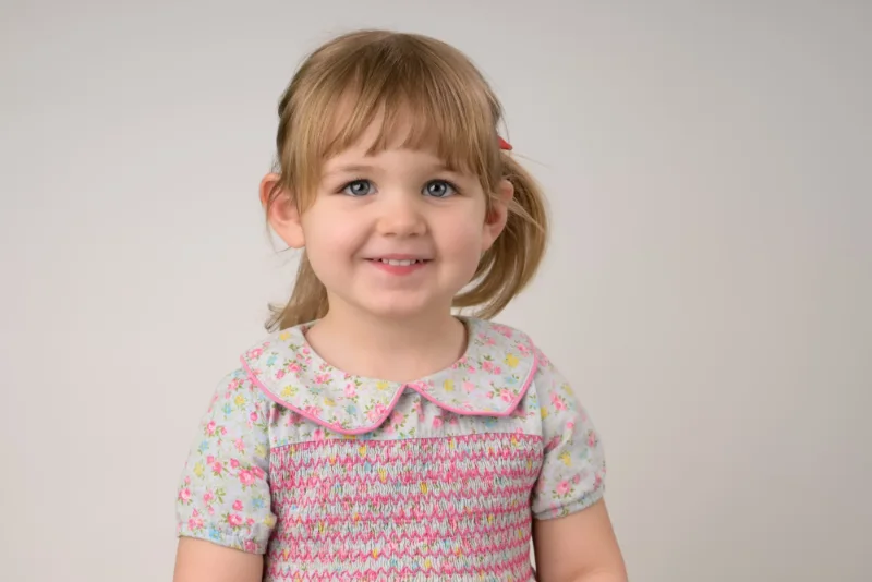 A toddler girl in a floral smocked dress smiling at the start of her photo session