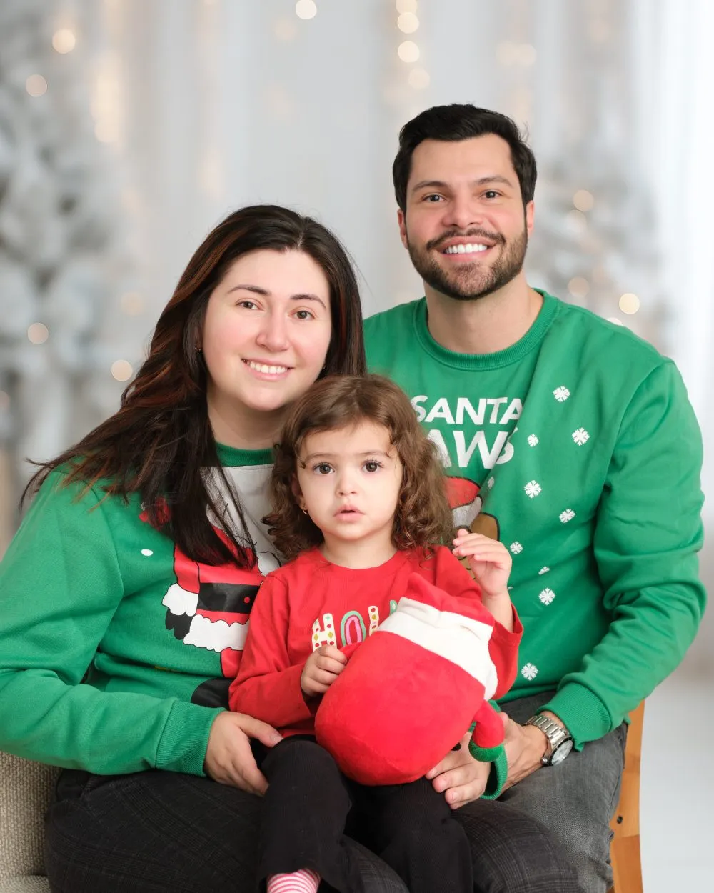 Family of three in green holiday sweaters in front of warm holiday lights