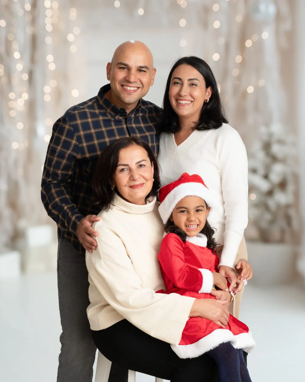 Three generations together with a child in a Santa dress in front of warm holiday lights