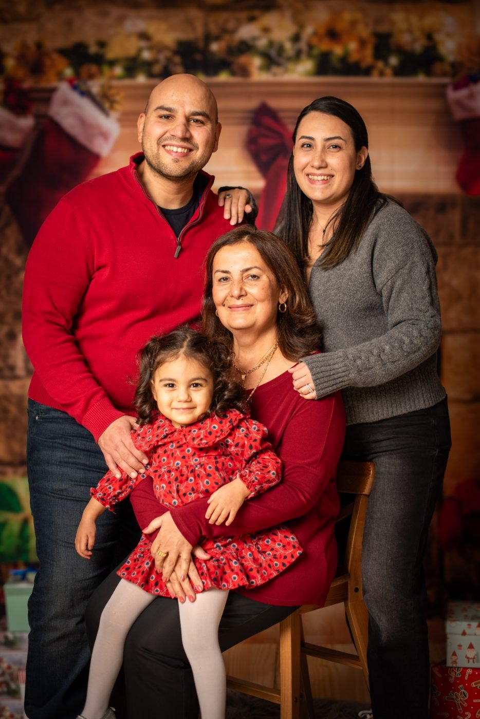 Three generations of a family in red and grey sweaters with a young girl on her grandmother's lap in front of a fireplace with stockings