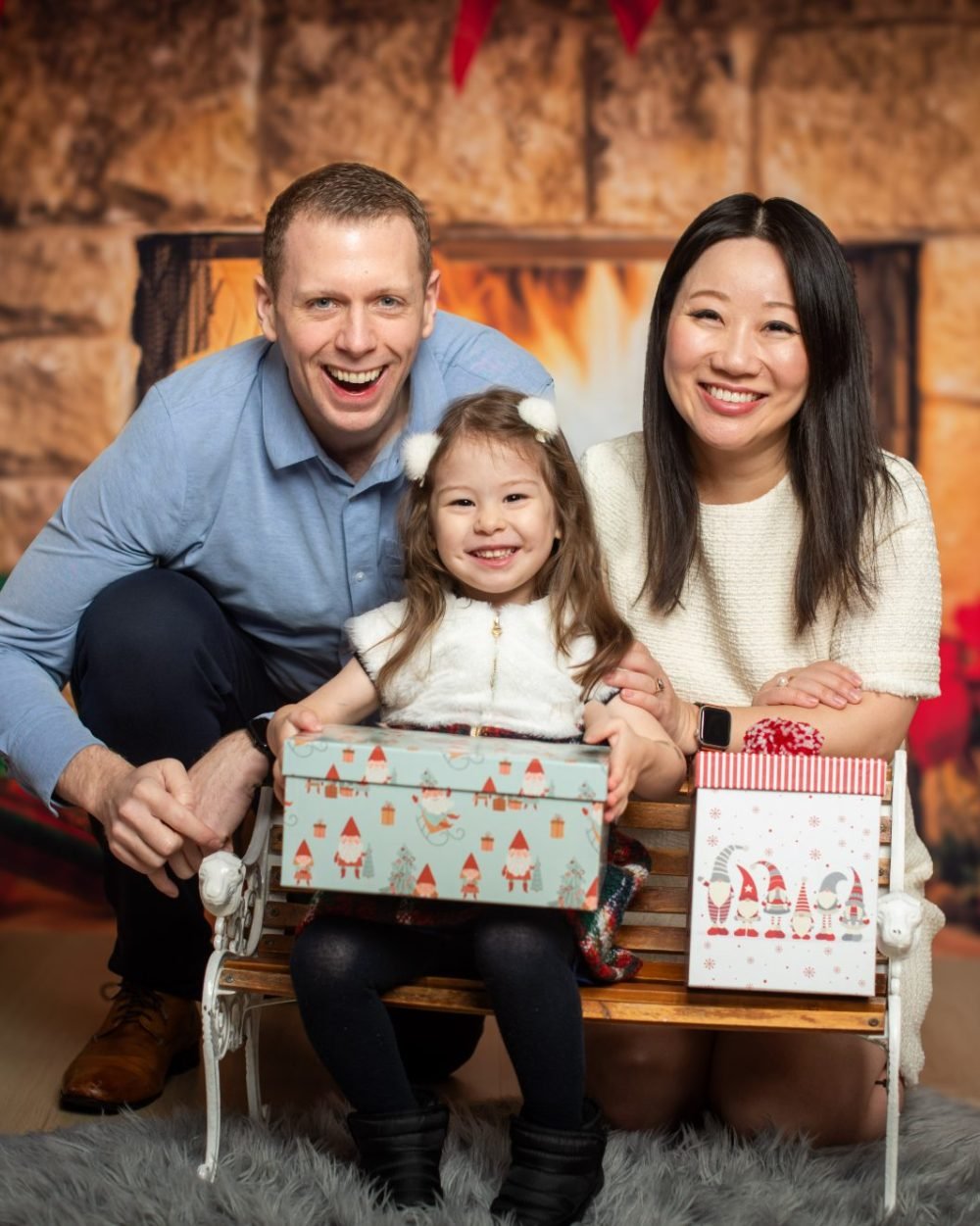 Parents and their young daughter holding wrapped gifts on a bench in front of a fireplace set