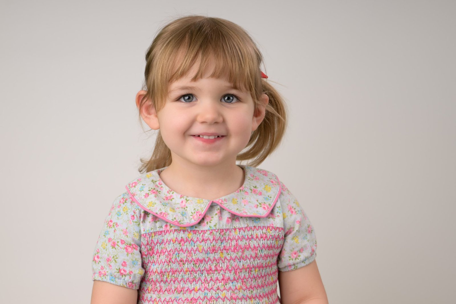 A toddler girl in a floral smocked dress smiling at the start of her photo session