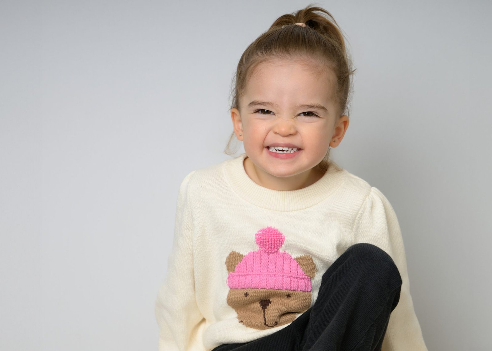A toddler girl in a cream bear-motif sweater with a scrunched-nose grin
