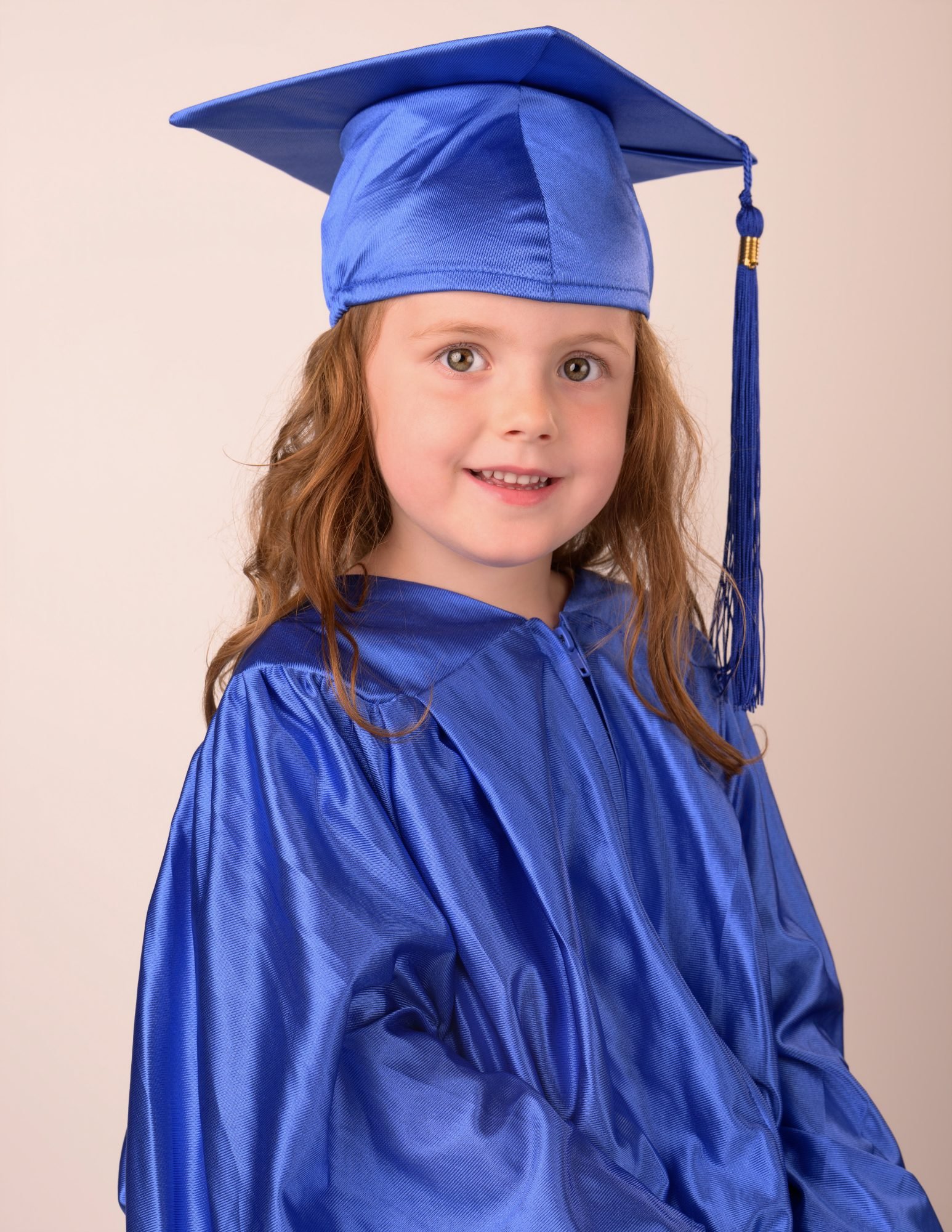 A preschool graduate with auburn hair smiling in her blue cap and gown