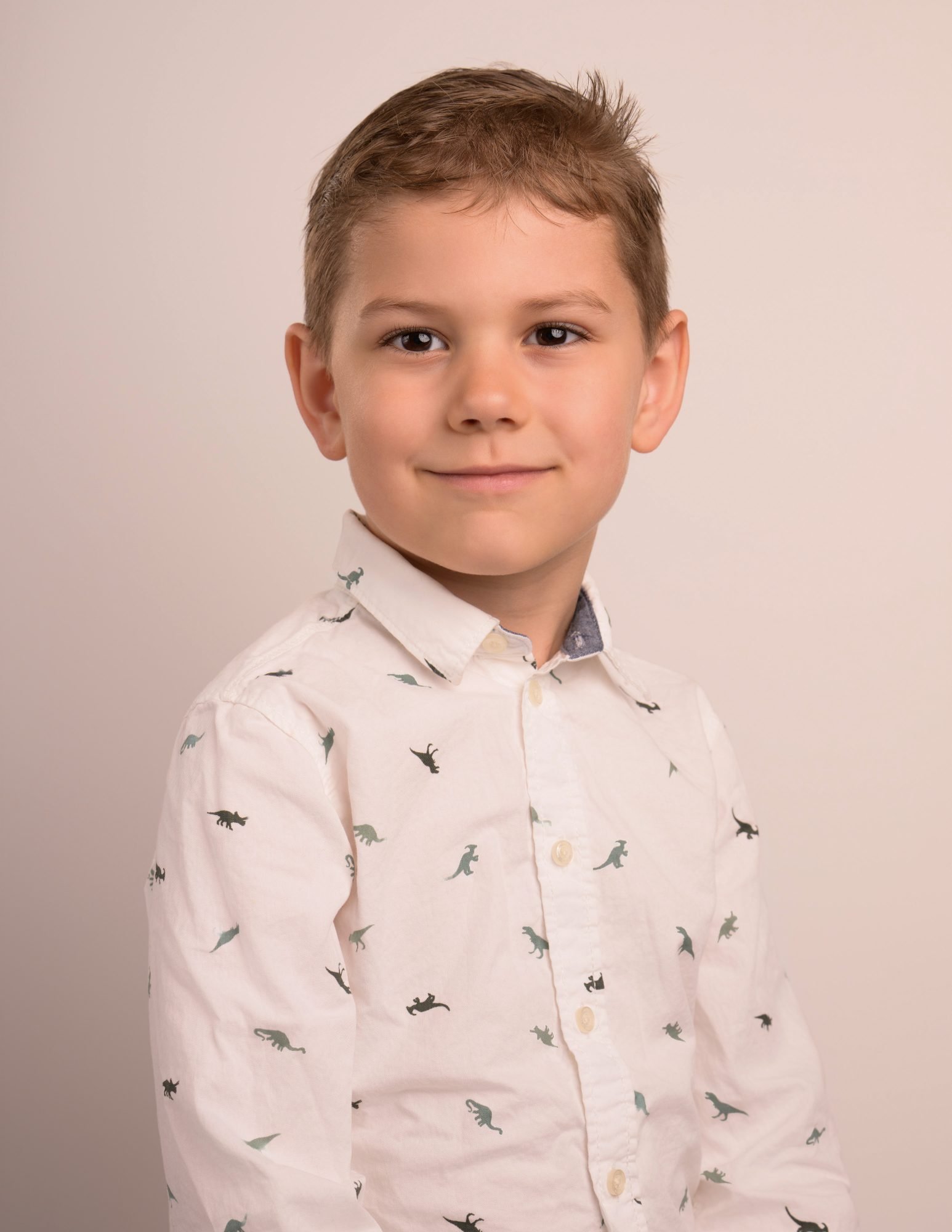 A young boy in a dinosaur-print shirt looking straight to camera on a warm neutral backdrop