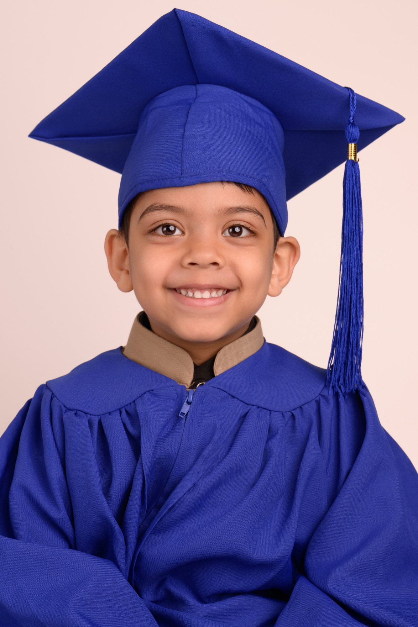 A preschool graduate in a blue cap and gown smiling on a warm blush backdrop
