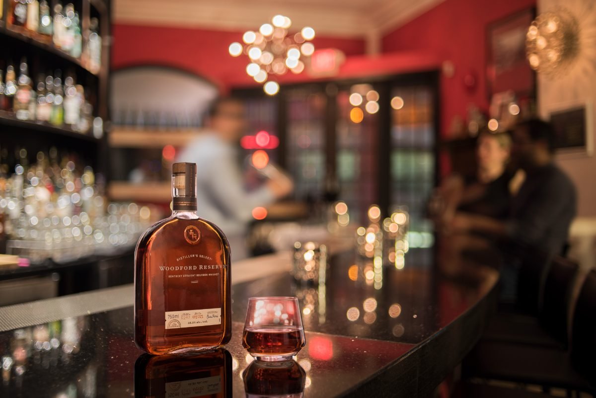 Beverage photography of a bourbon bottle and poured glass reflected on a dark cocktail bar
