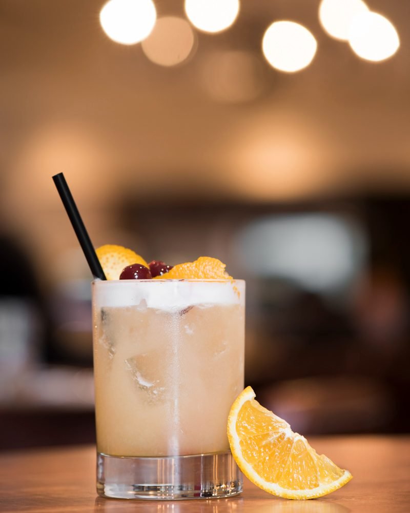 Cocktail photography of an amaretto sour with orange and cherry garnish on a warm-lit Vancouver bar