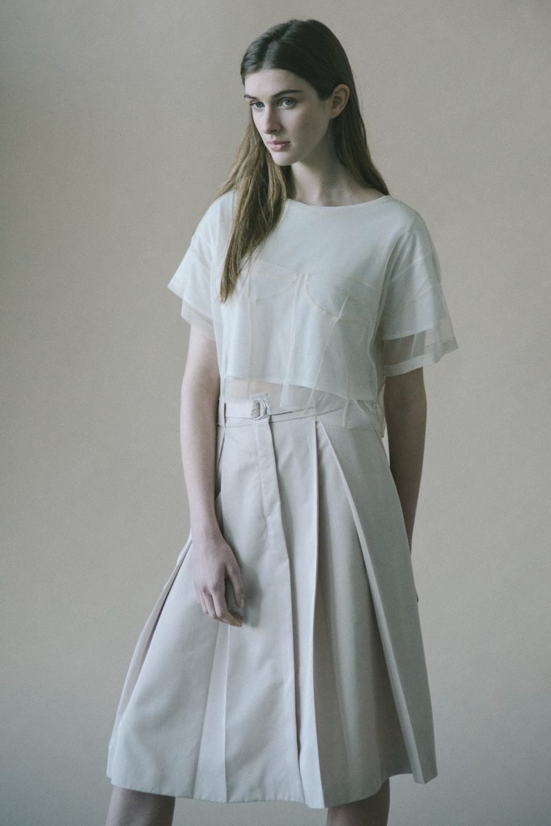Fashion lookbook photography of a model in a sheer cream top and pleated skirt on a warm studio backdrop