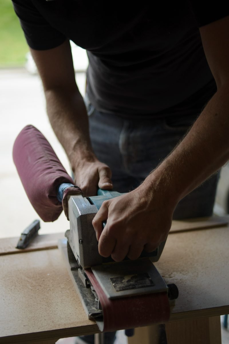 Close-up manufacturing photography of a tradesperson's hands running a belt sander over a hardwood edge