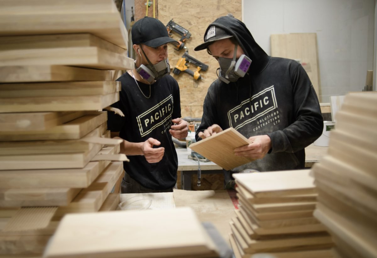 Industrial photography of two millwork tradesmen in respirators inspecting a finished maple panel in a Lower Mainland cabinet shop