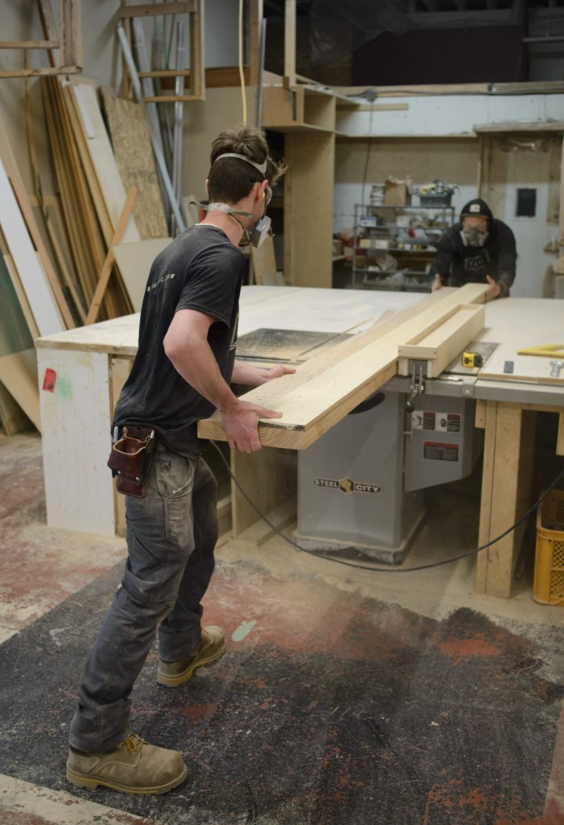 Manufacturing photography of two workers feeding a long panel through a table saw in a fabrication shop