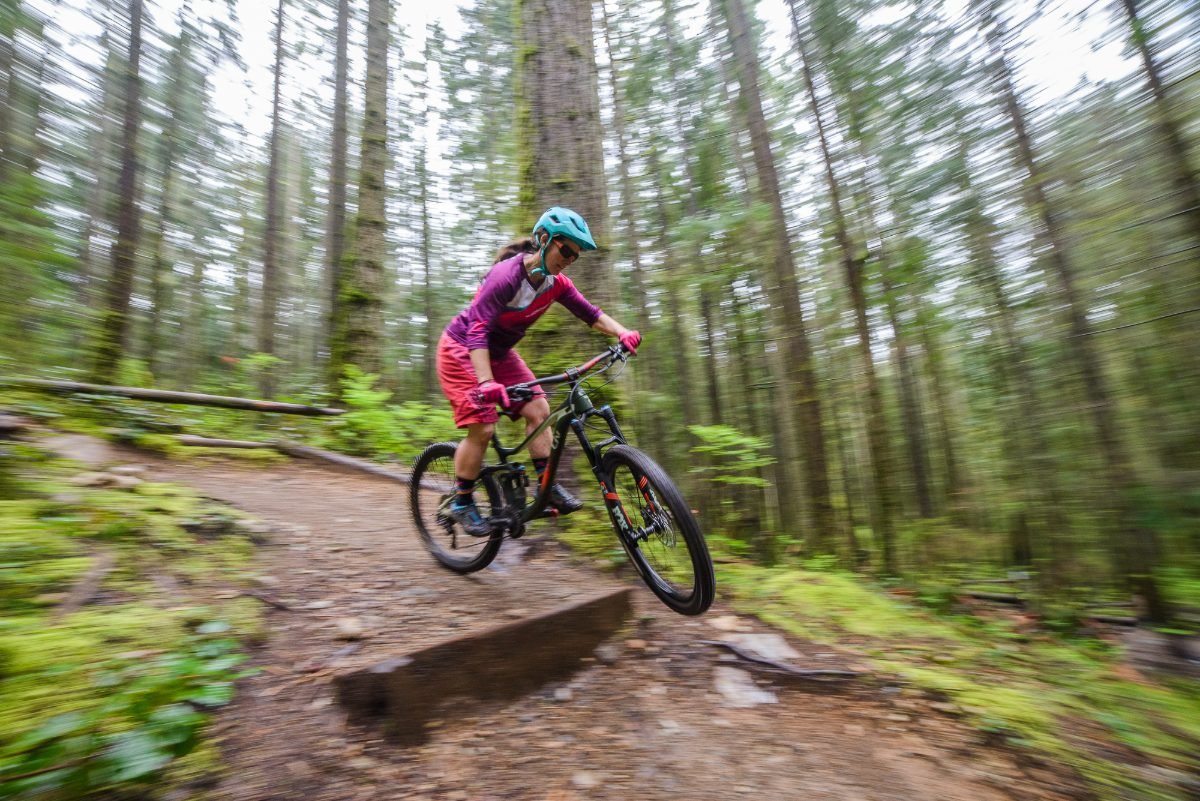 Outdoor brand photography of a mountain biker launching a trail feature with motion-blurred forest behind