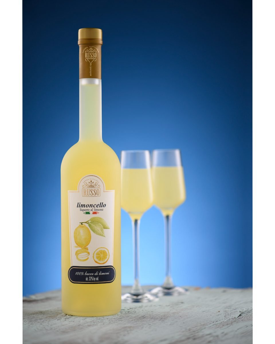 Product photography of a limoncello bottle and two chilled glasses on a blue studio gradient