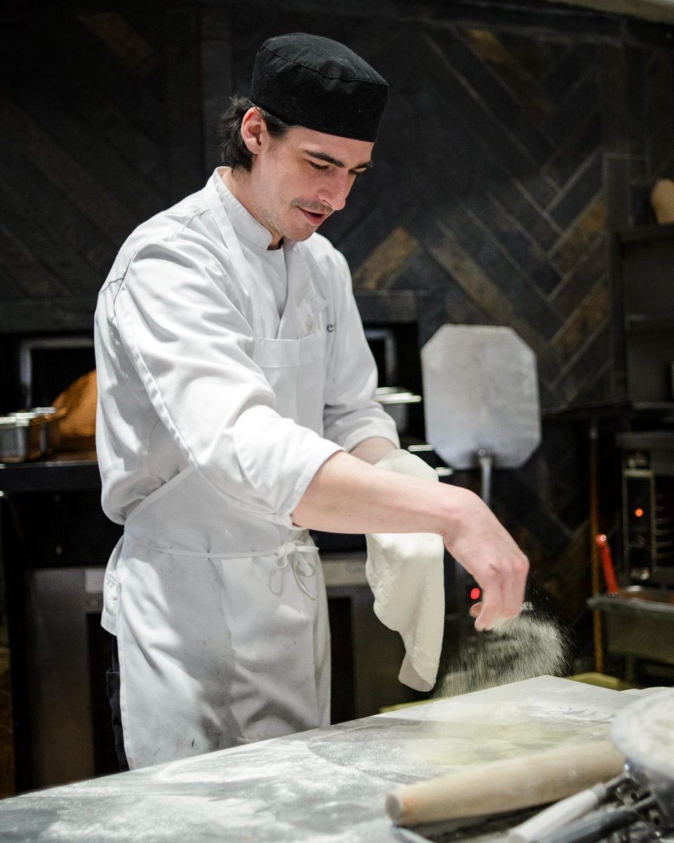 Restaurant photography of a chef dusting flour over pizza dough beside a wood-fired oven