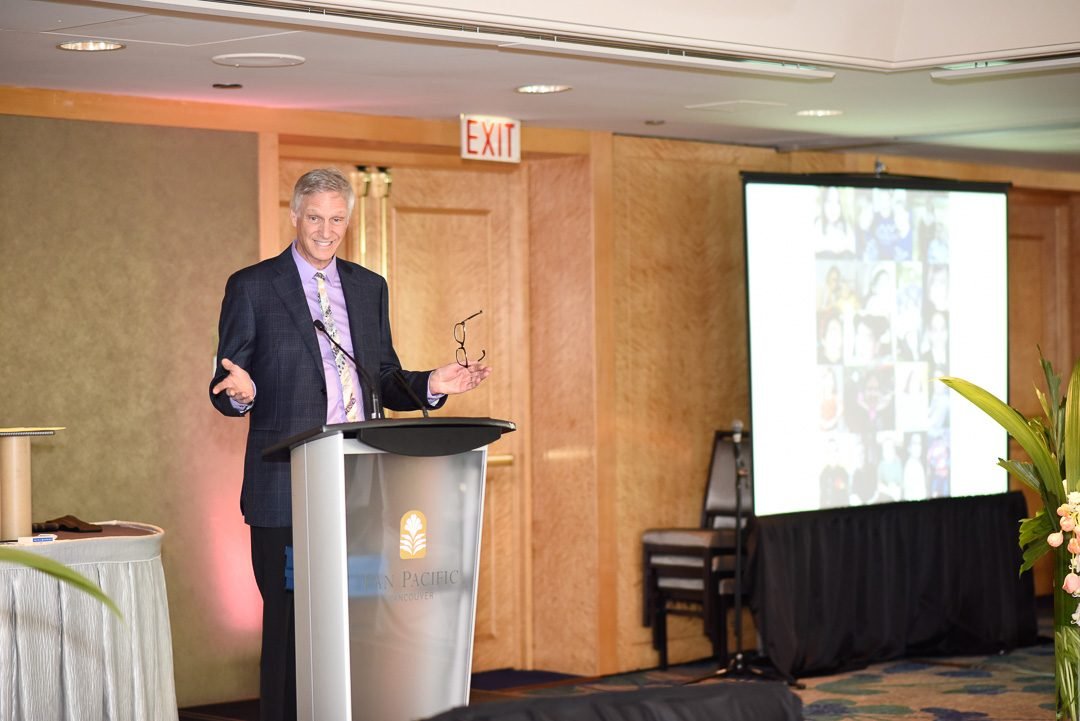 Master of ceremonies speaking from the podium during a fundraising gala in a downtown Vancouver hotel ballroom
