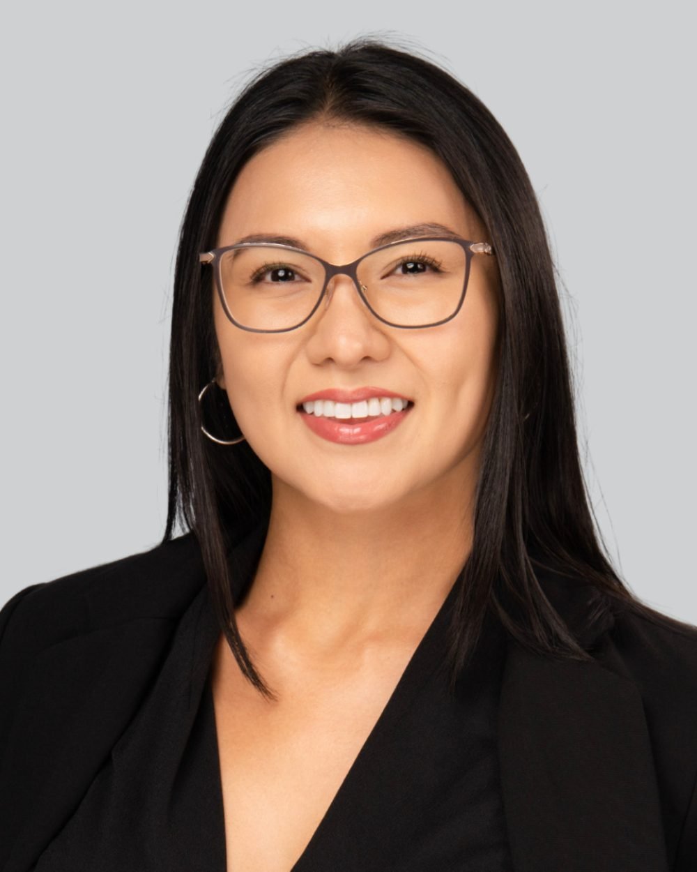 Event headshot booth portrait of a smiling woman in a black blazer and glasses on a grey backdrop