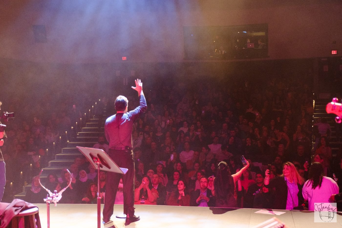 Concert performer photographed from behind on stage as the audience raises their hands