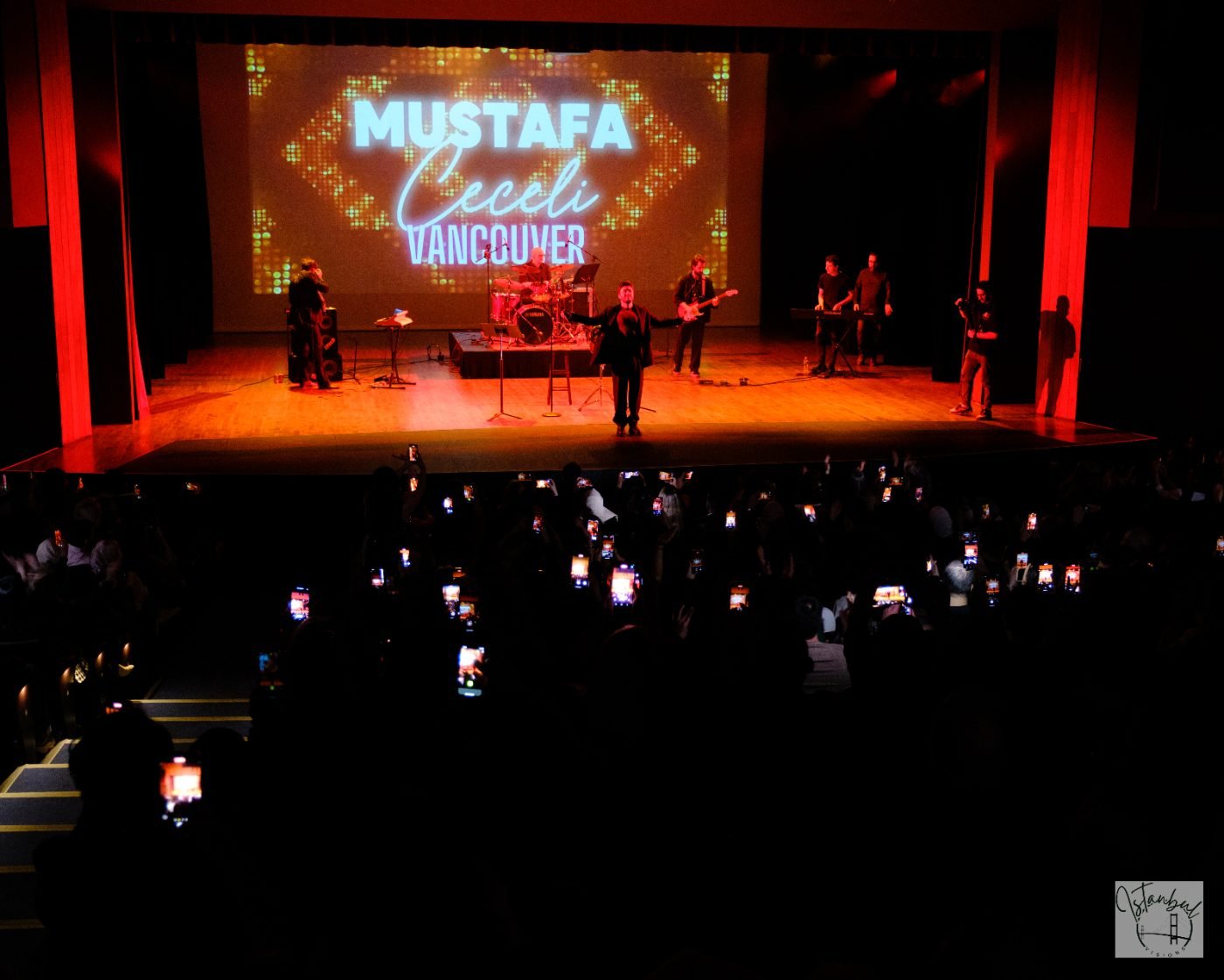 Performer standing arms open on stage while the audience films with their phones at a theatre concert in Vancouver