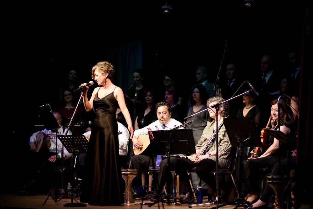 Live music photography of a singer performing with a seated ensemble behind her on a concert stage