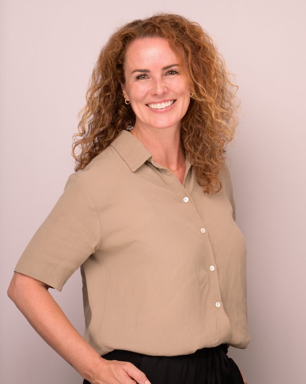 Professional headshot of a smiling woman in a tan shirt on a soft pink studio backdrop