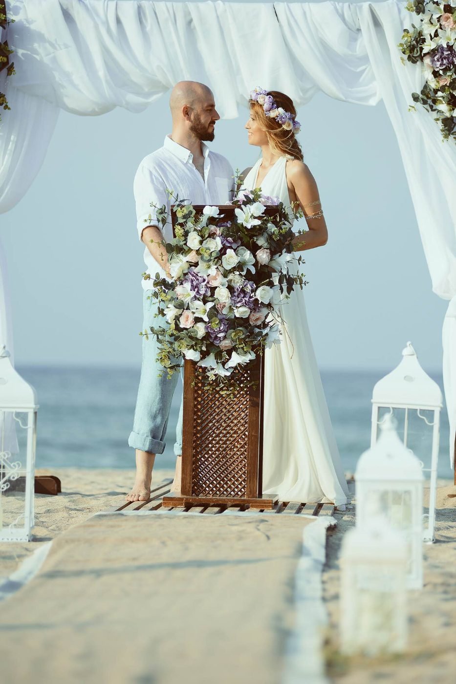 Beach wedding ceremony photographed at a floral podium under a white draped arch at the water&rsquo;s edge
