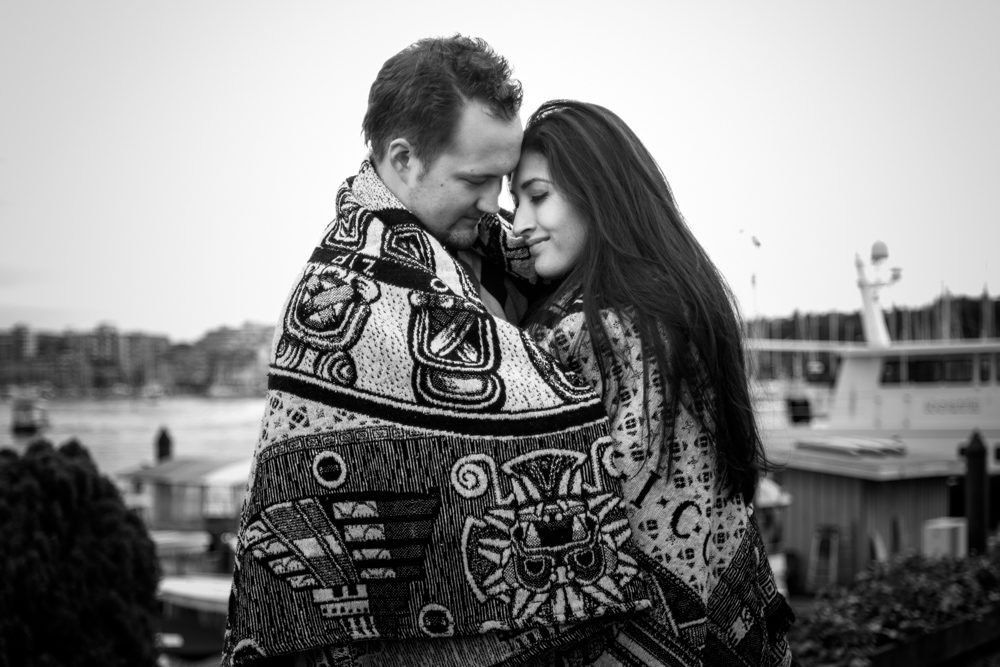 Black and white engagement portrait of a couple wrapped in a blanket at the waterfront