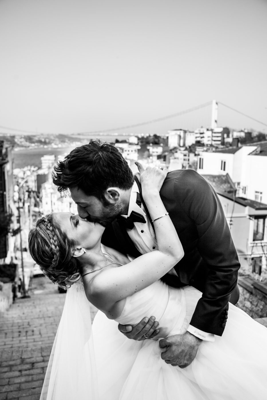 Black and white wedding portrait of the groom dipping the bride for a kiss on a hillside street above the water