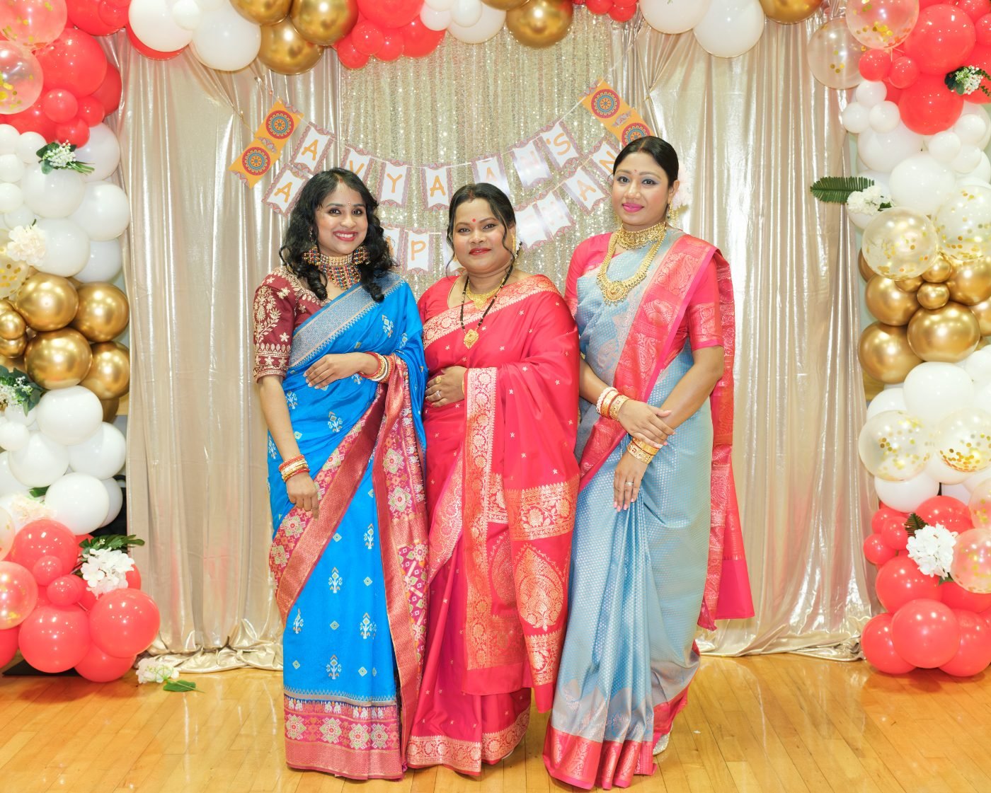 Three guests in bright saris photographed together in front of a balloon arch at a family celebration in the Lower Mainland