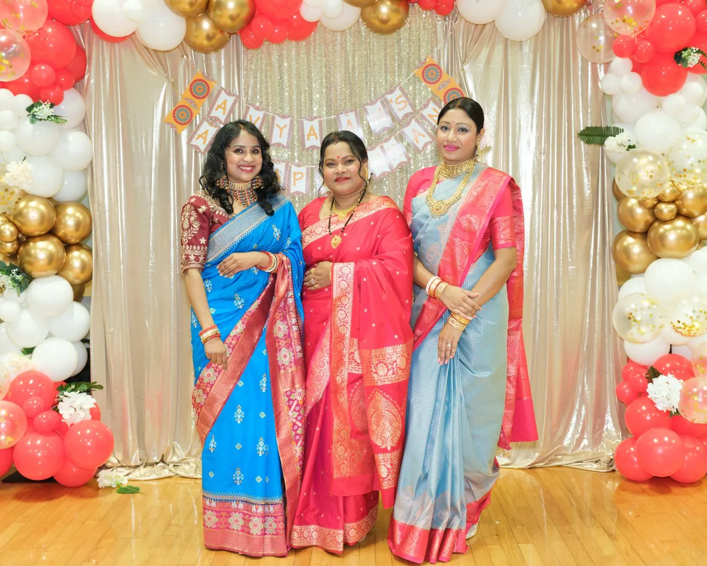 Three guests in bright saris photographed together in front of a balloon arch at a family celebration in the Lower Mainland