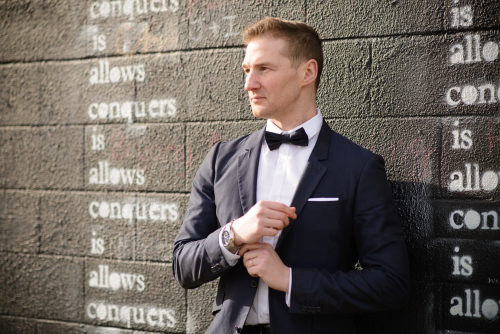 Groom portrait in a navy suit and black bow tie against a painted brick wall on the morning of the wedding
